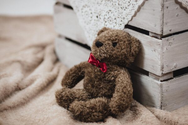 Charming teddy bear with a red bowtie, resting against a rustic wooden crate on a soft, cozy blanket.