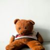 Cute brown teddy bear wearing a sweater, sitting on a white floor against a plain background.